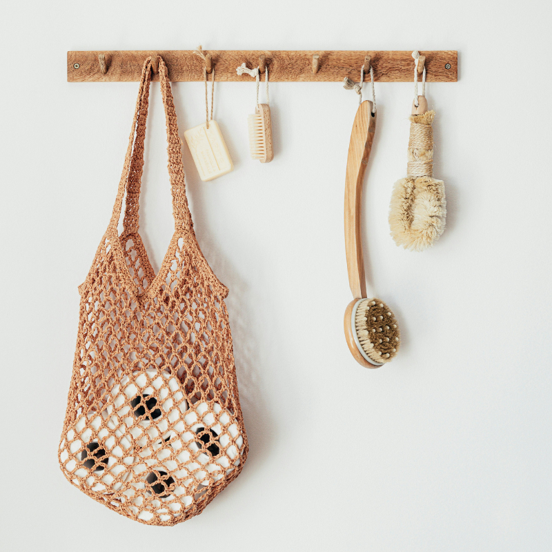 Plastic-free bathroom accessories on a wooden rack, including a natural fibre bag, bamboo brushes, and soap pouch.