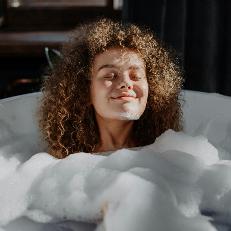Smiling woman with curly hair enjoying a relaxing bubble bath, symbolising self-care and relaxation.