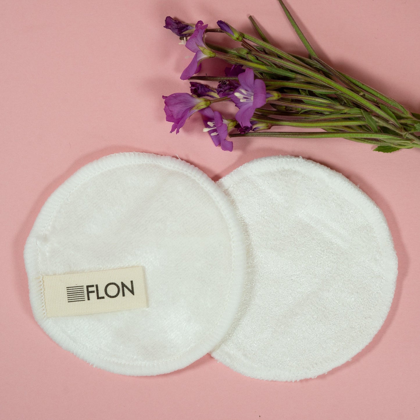 Two reusable bamboo makeup remover pads with a small bouquet of purple flowers on a pink background.