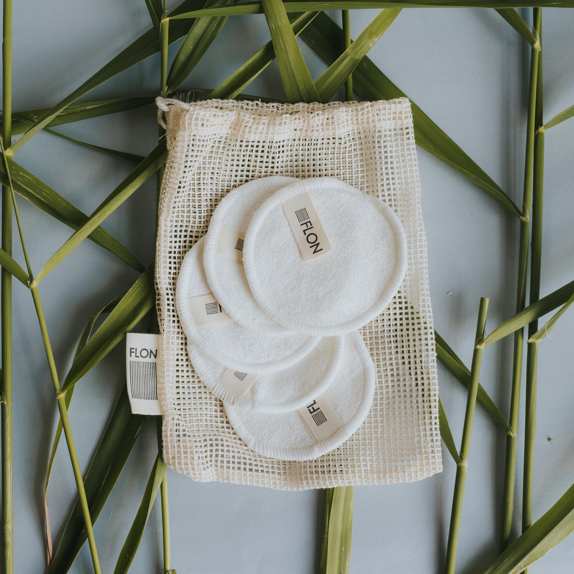 Set of reusable bamboo makeup remover pads stacked with mesh wash bag on a green leafy background.