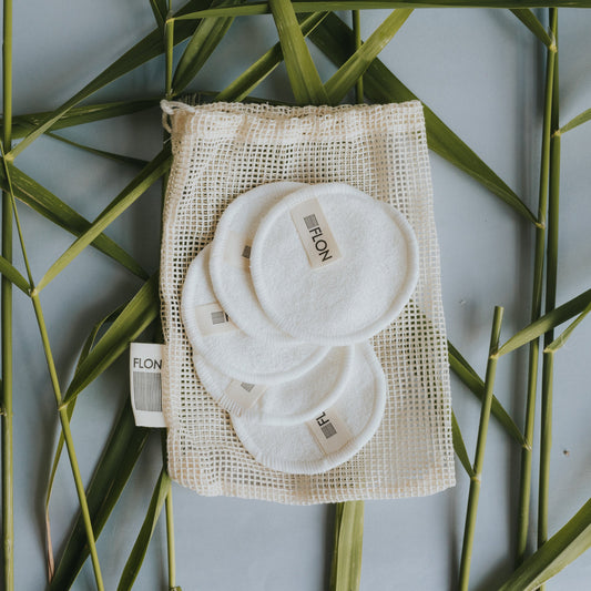 Set of reusable bamboo makeup remover pads stacked with mesh wash bag on a green leafy background.